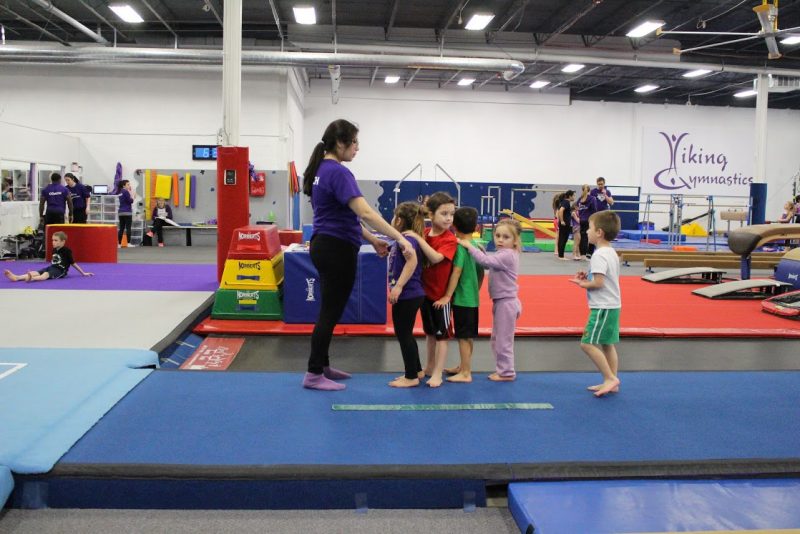 A gymnastics coach leads a line of five young children across a colorful gym.