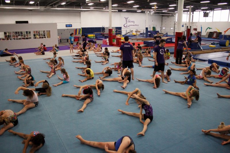 A group of young gymnasts stretches in a straddle position on a gymnastics floor, overseen by a number of purple-clad Viking coaches.