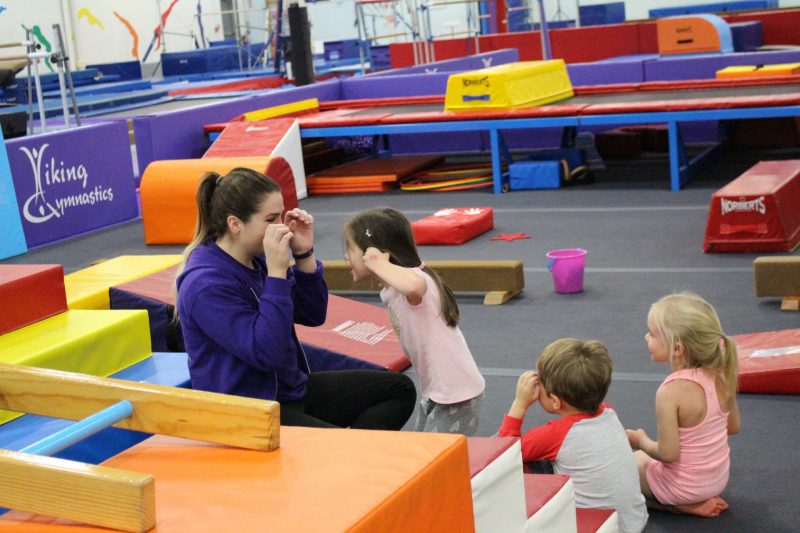 A gymnastics coach talks to a class of three toddler-aged children.