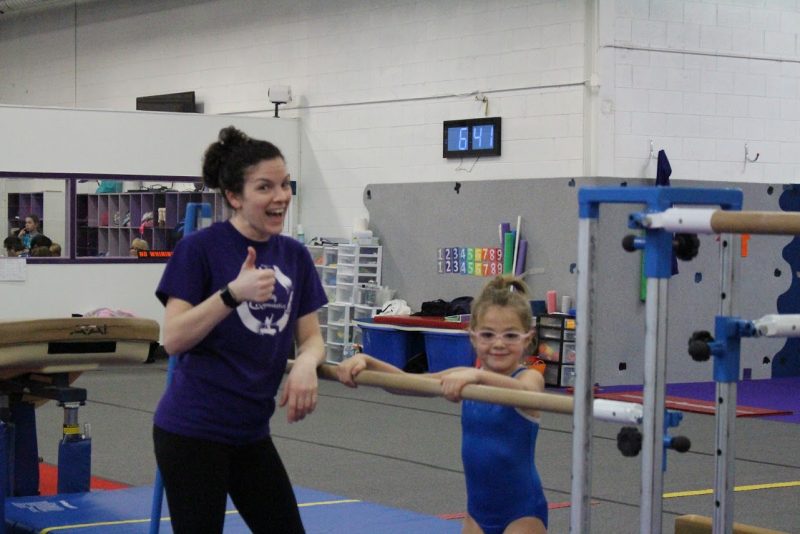 A purple-clad gymnastics coach gives a big thumbs up and a smile alongside her bespectacled gymnasts on the bars.