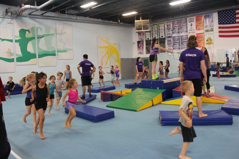 A photograph of children running around a floor; a pile of gymnastics mats is stacked in the middle of the floor as part of a game the children are playing.